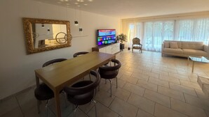 Un salon moderne avec un canapé, un coin repas comprenant une table et des chaises, un téléviseur et une grande fenêtre avec des rideaux.