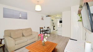 A living room with a sofa, coffee table with flowers, a TV, and a kitchen in the background. - Stylish & Comfy | Near Canary Wharf | Self Check-In | WiFi (London)