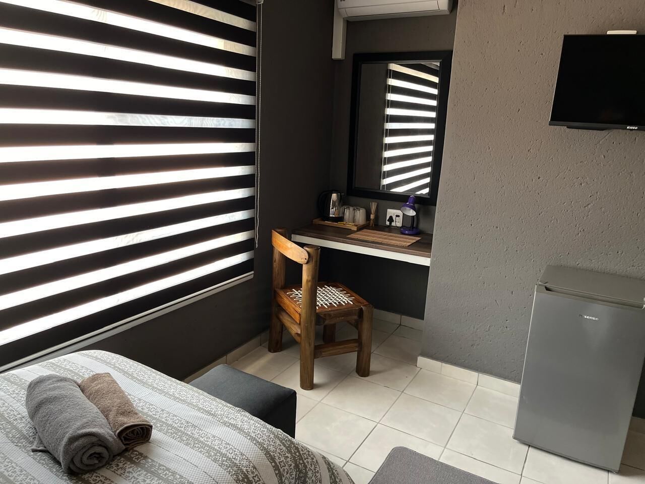 Habitació petita i moderna amb un llit, una cadira de fusta, escriptori amb mirall i un frigorífic.