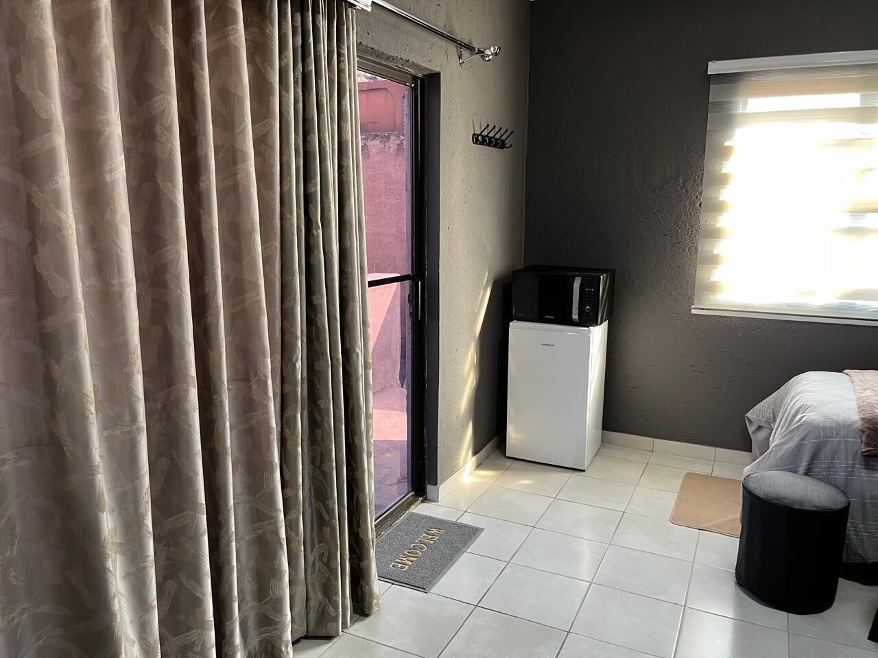 A room with a bed, a small white appliance, a window with blinds, and a door with curtains.