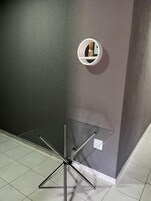 A corner of a room with a grey textured wall, a small white shelf with a mirror, and a metal-framed table with a reflective surface.