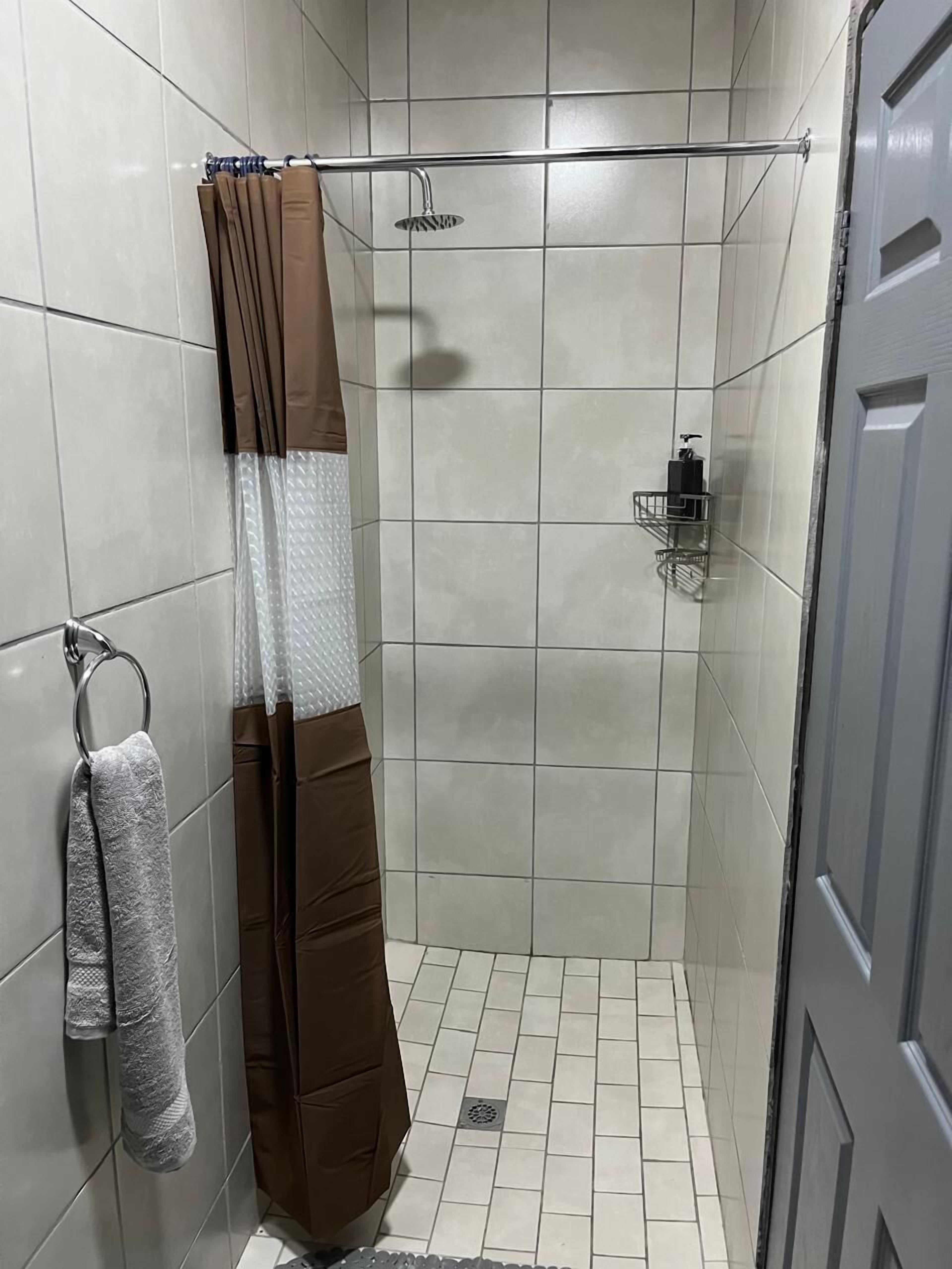 A small, tiled shower area with a showerhead, a towel rack with a towel, and a soap dispenser on the wall.