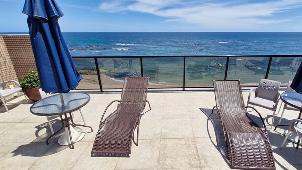 Un balcón con dos sillones, una mesa y una sombrilla, con vista al océano.