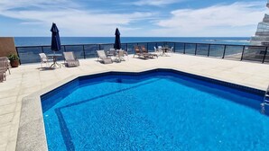 Una piscina en la azotea con vista al océano, rodeada de sillas de descanso y sombrillas.
