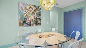 A dining area with a glass table surrounded by chairs, a painting on the wall, and a blue door. - Blue Sky Day Beach Cottage (Anna Maria)