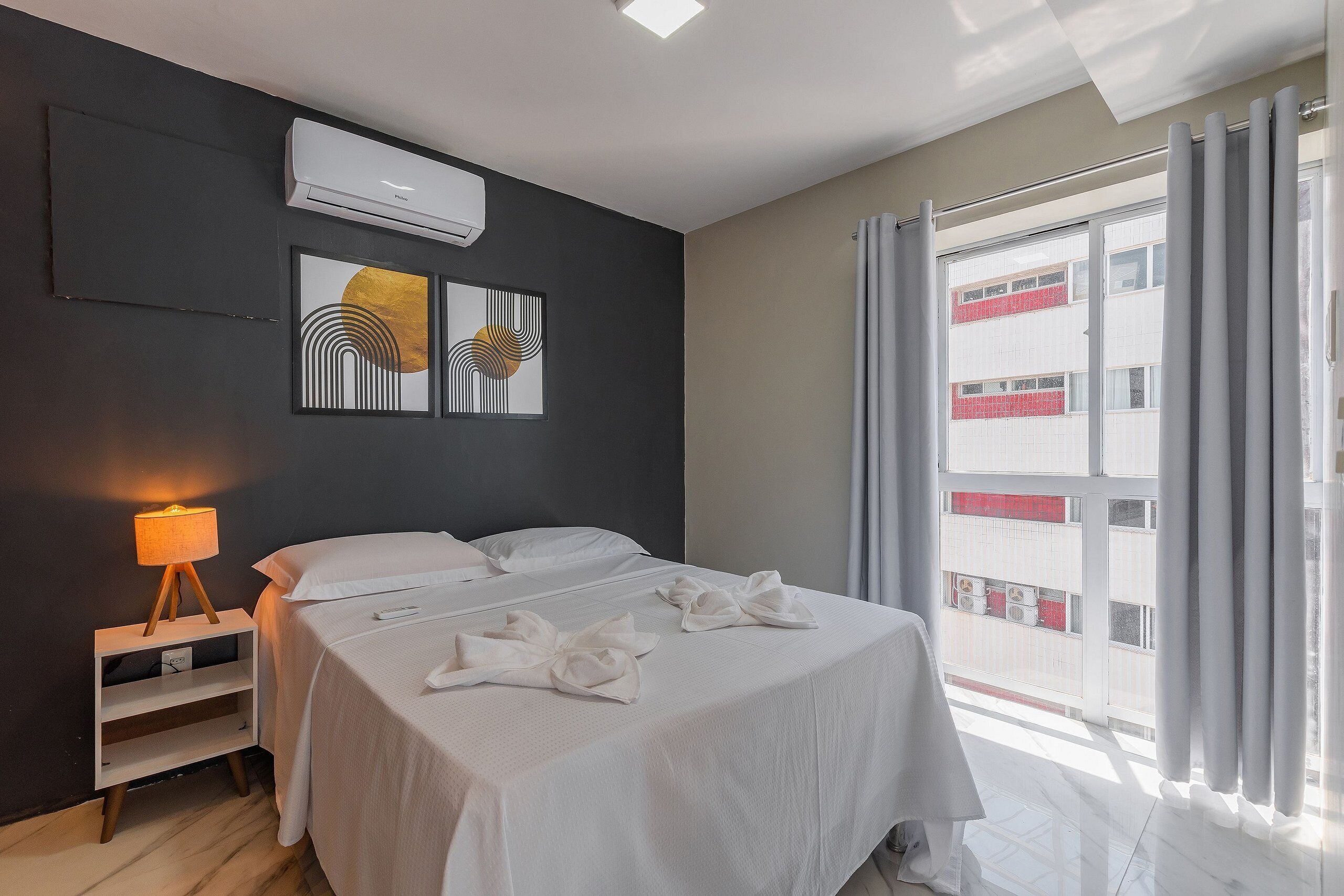 A hotel room with a bed, a bedside table with a lamp, a wall-mounted air conditioner, and a window with curtains.