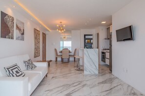 Ein modernes Wohnzimmer mit einem weißen Sofa, einem Essbereich mit Tisch und Stühlen, einer Küchenzeile mit an der Wand montiertem Fernseher und einem Flur, der in einen weiteren Raum führt.