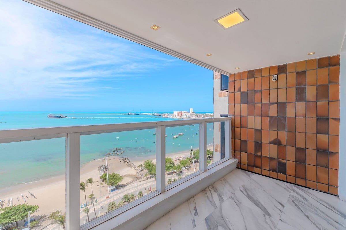 A balcony with a view of the beach and ocean, a tiled wall, and a marble floor.