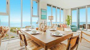 Une vaste salle à manger avec une grande table en bois, dressée pour six personnes, entourée de fauteuils en cuir. La pièce est dotée de grandes fenêtres offrant une vue sur l’océan et le panorama de la ville.