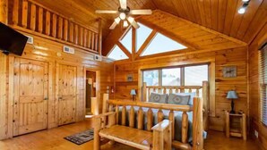 L’intérieur d’une cabane en rondins, avec un banc en bois, un ventilateur de plafond et une fenêtre avec des stores.