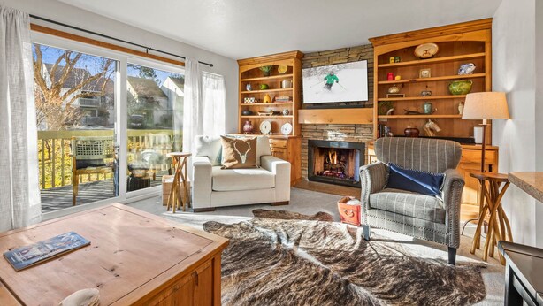 A cozy living room with a fireplace, a television, and a large window overlooking a garden. - Moose Mgmt – Pvt Hot Tub | Free Shuttle | Sleeps 8 (Park City)