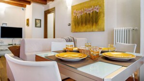 Un comedor con una mesa preparada para cuatro personas, sillas blancas, un cuadro en la pared y un televisor al fondo.