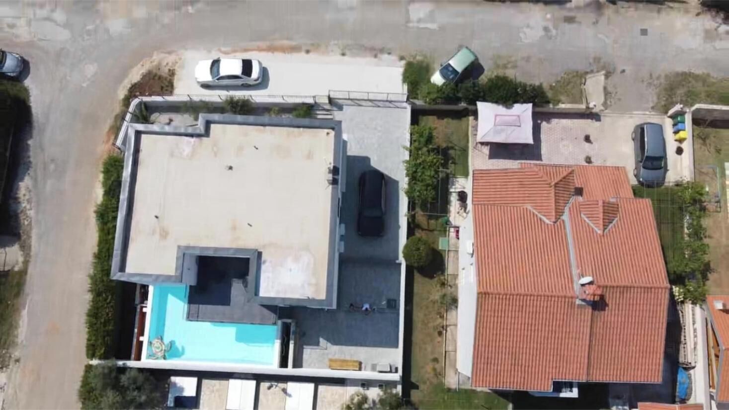 Vista aérea de una zona residencial con una casa que tiene piscina, un edificio de techo rojo y varios coches estacionados.