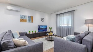 Ein modernes Wohnzimmer mit grauen Sofas, einem Flachbildfernseher, einem Couchtisch und einem großen Fenster mit durchscheinenden Vorhängen.