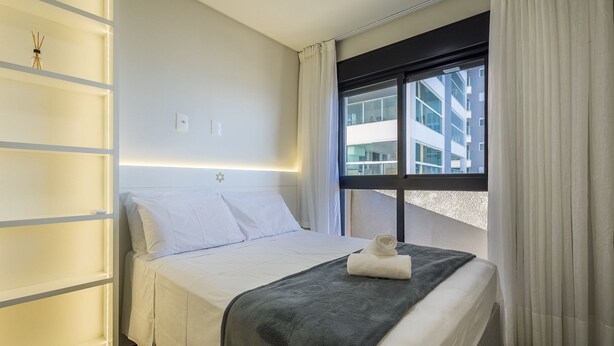 Una camera d'albergo con un letto, una finestra con tende, uno scaffale a muro con una bottiglia e una decorazione a Stella di David.