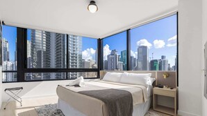 A modern bedroom with a large bed, a nightstand, and a view of a city skyline through large windows. - 1 BR | Rooftop Pool Deck & Co-Working Floor (Miami)