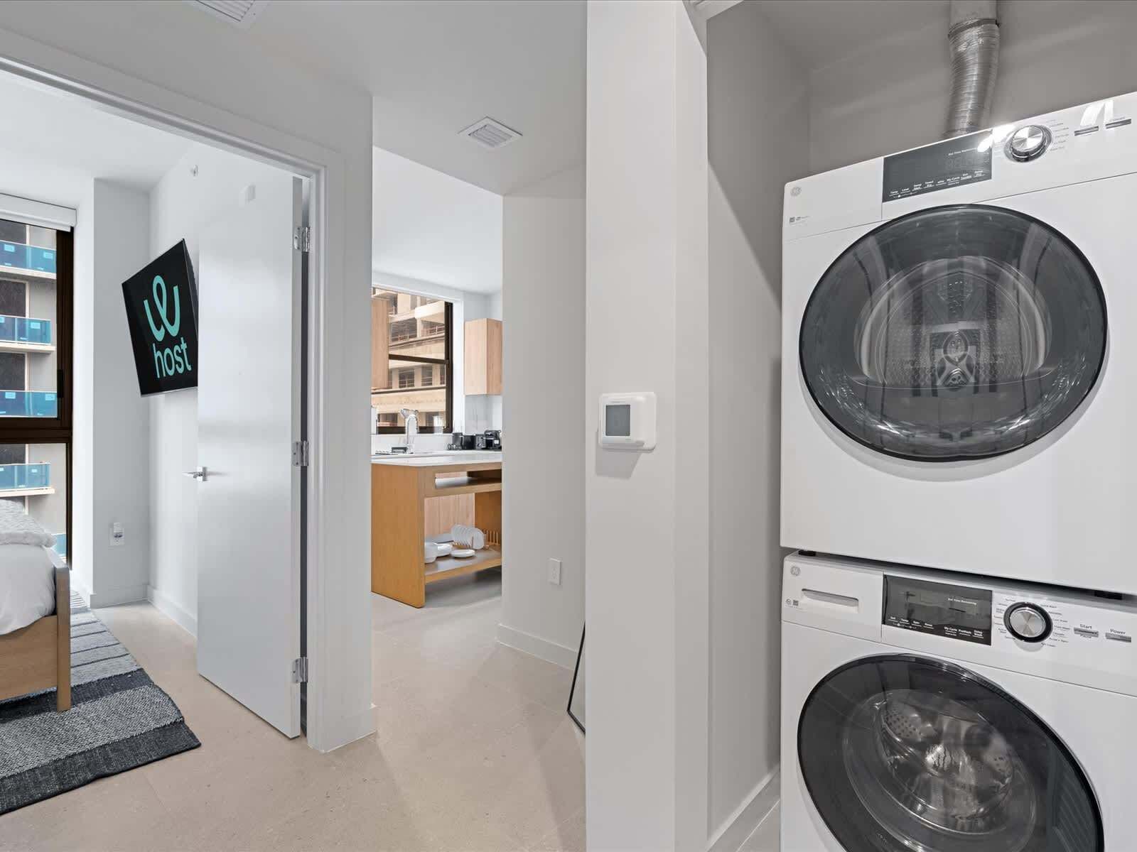 A laundry room with two stacked front-loading washing machines and a small kitchen area visible through an open door.