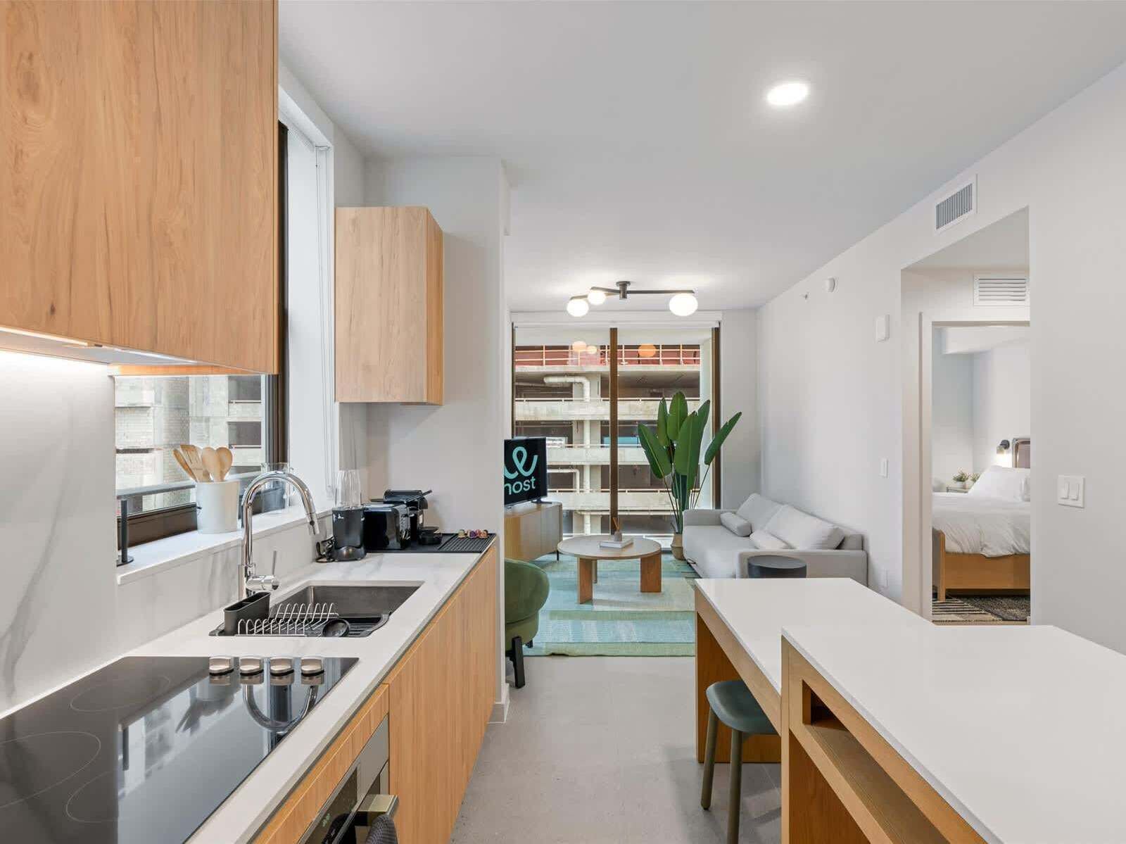 A modern kitchen with a built-in oven, a sink, and a coffee machine. There is a dining area with a table and chairs, and a living room with a sofa and a plant.