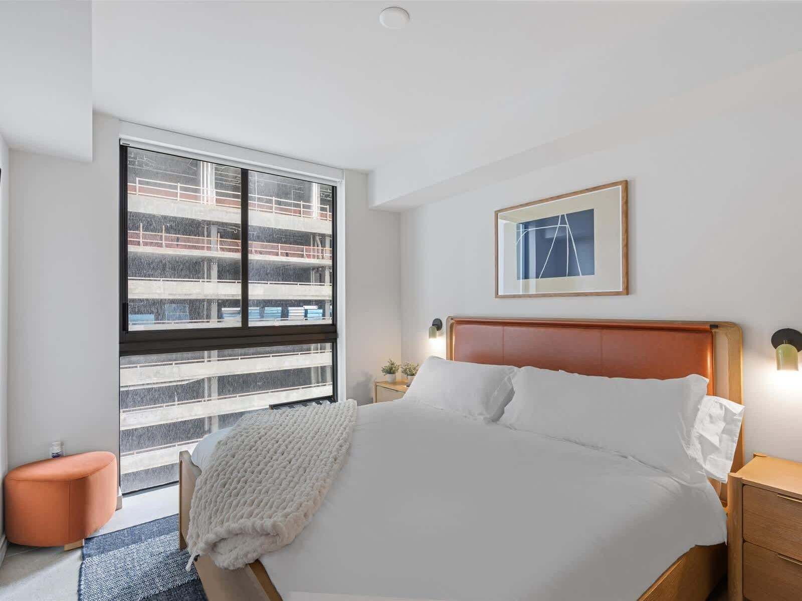 A hotel room with a large window, a bed with white linens, a wooden headboard, a nightstand with a lamp, and a framed picture on the wall.