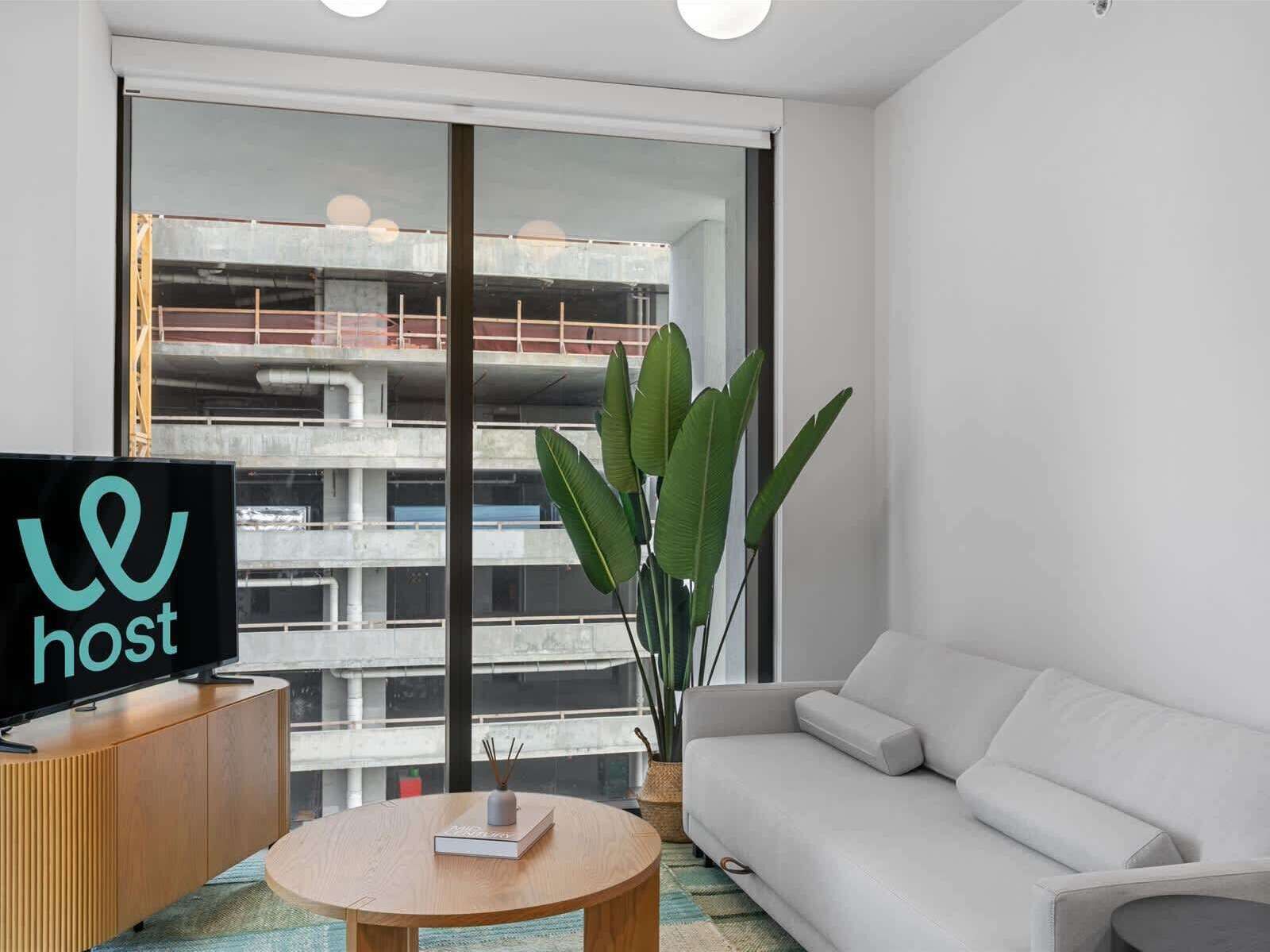 A modern living room with a sofa, a coffee table, and a television on a wooden cabinet. There is a large window with a view of a building under construction.