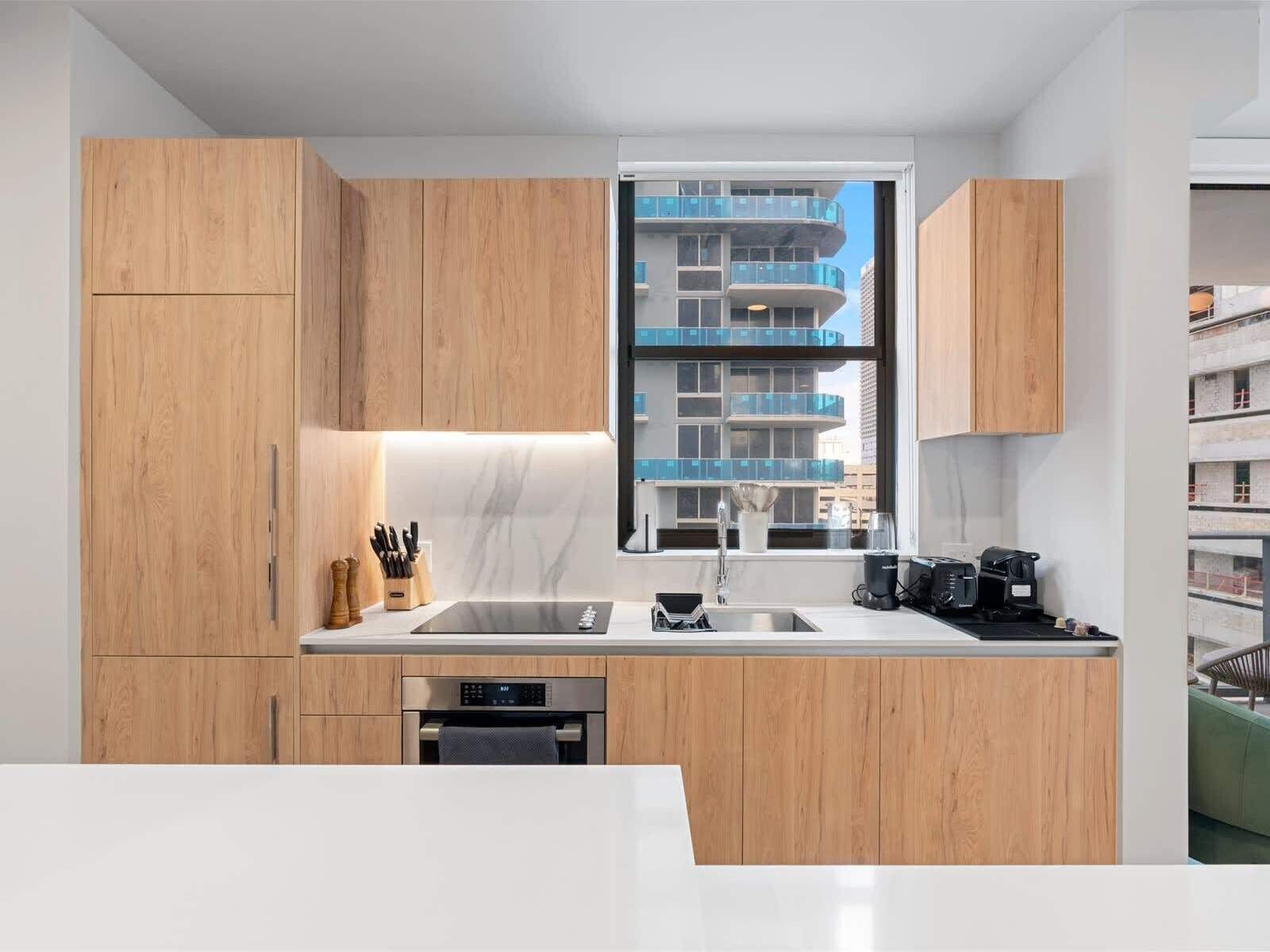 Une cuisine moderne avec des meubles en bois, un évier en acier inoxydable et une fenêtre donnant sur un immeuble à plusieurs étages.