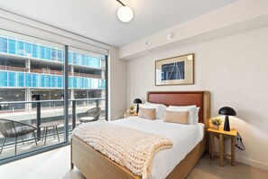 Una habitación de hotel moderna con una cama grande, una mesita de noche con lámpara, una silla y vistas a un edificio a través de amplios ventanales.
