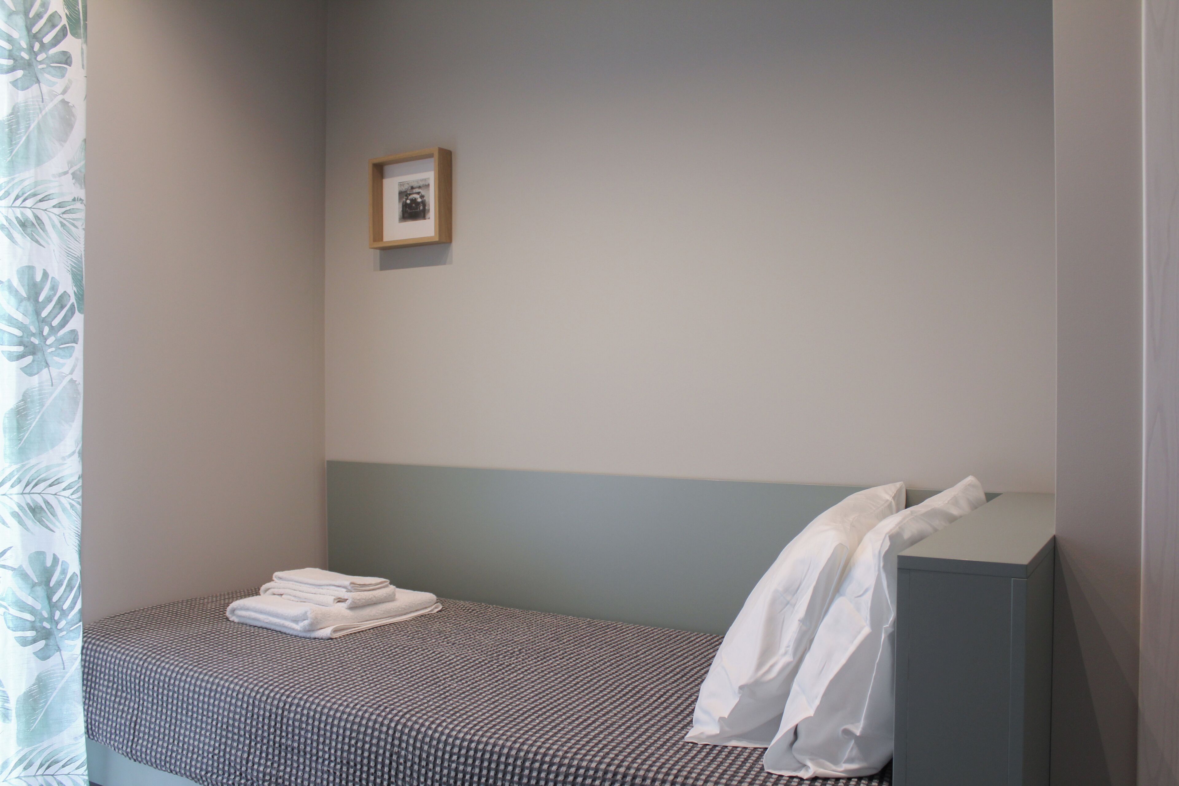 Ein kleines Zimmer mit einem Einzelbett, einer karierten Bettdecke, einem ordentlich gefalteten weißen Handtuch und einem weißen Hemd, das über der Kopfteil drapiert ist.