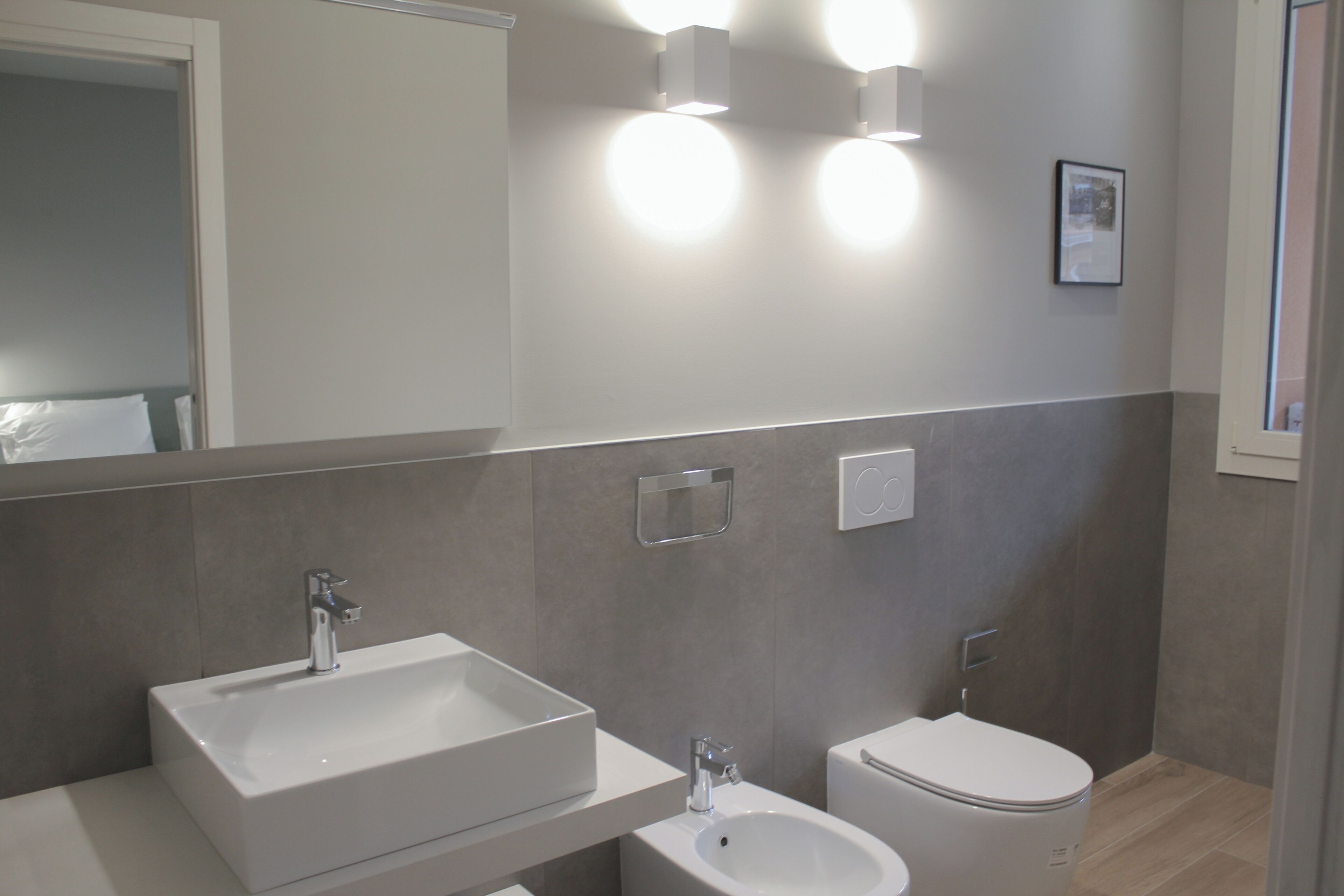 A modern bathroom with a rectangular sink, a bidet, and a toilet. There are wall-mounted lights and a framed picture on the wall.