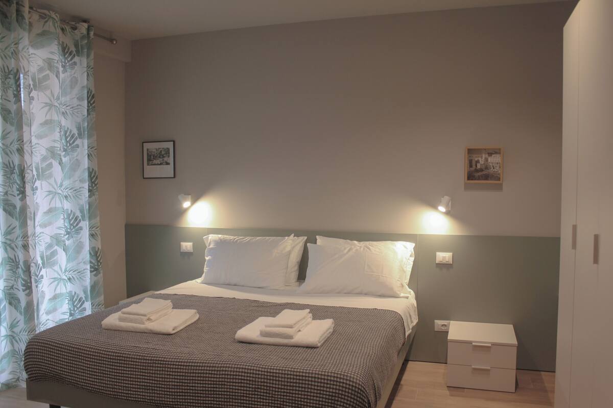 Un lit bien fait, avec du linge de lit blanc et une couvre-lit à carreaux, une table de chevet avec une lampe, et une petite table de nuit avec un tiroir.