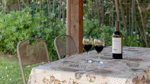 Une table avec une bouteille de vin et deux verres de vin rouge, disposée à l’extérieur, avec une chaise et de la verdure en arrière-plan.