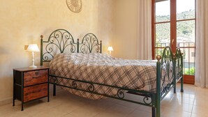 Une chambre à coucher avec un lit en métal, deux tables de chevet avec des lampes, une couverture à carreaux, une horloge murale et une fenêtre avec des rideaux.