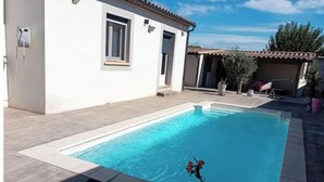 Une piscine rectangulaire avec une terrasse carrelée, un bâtiment blanc doté d’un toit en tuiles et un pot de fleurs orné de fleurs rouges.