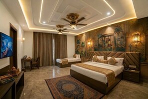 Une chambre d’hôtel avec un grand lit, deux petits lits, un ventilateur de plafond, une télévision et une fresque décorative sur un mur.