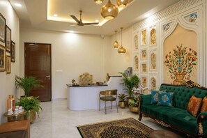 A well-lit living room with a green sofa, a coffee table, and a desk with a chair. There are decorative wall panels and a ceiling fan. - Ambassador Villa - An Experiential stay (Agra)
