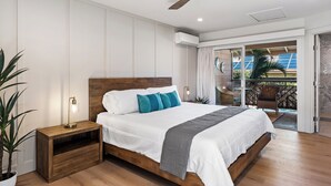 Ein modernes Schlafzimmer mit einem hölzernen Bett, weißer Bettwäsche, türkisfarbenen Kissen, einem Nachttisch mit Lampe und Blick auf einen Balkon mit Gartenmöbeln.
