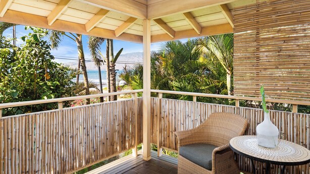 Un balcon abrité, agrémenté d’une chaise en osier et d’une petite table, donnant sur une plage bordée de palmiers et surmontée d’une montagne en arrière-plan.