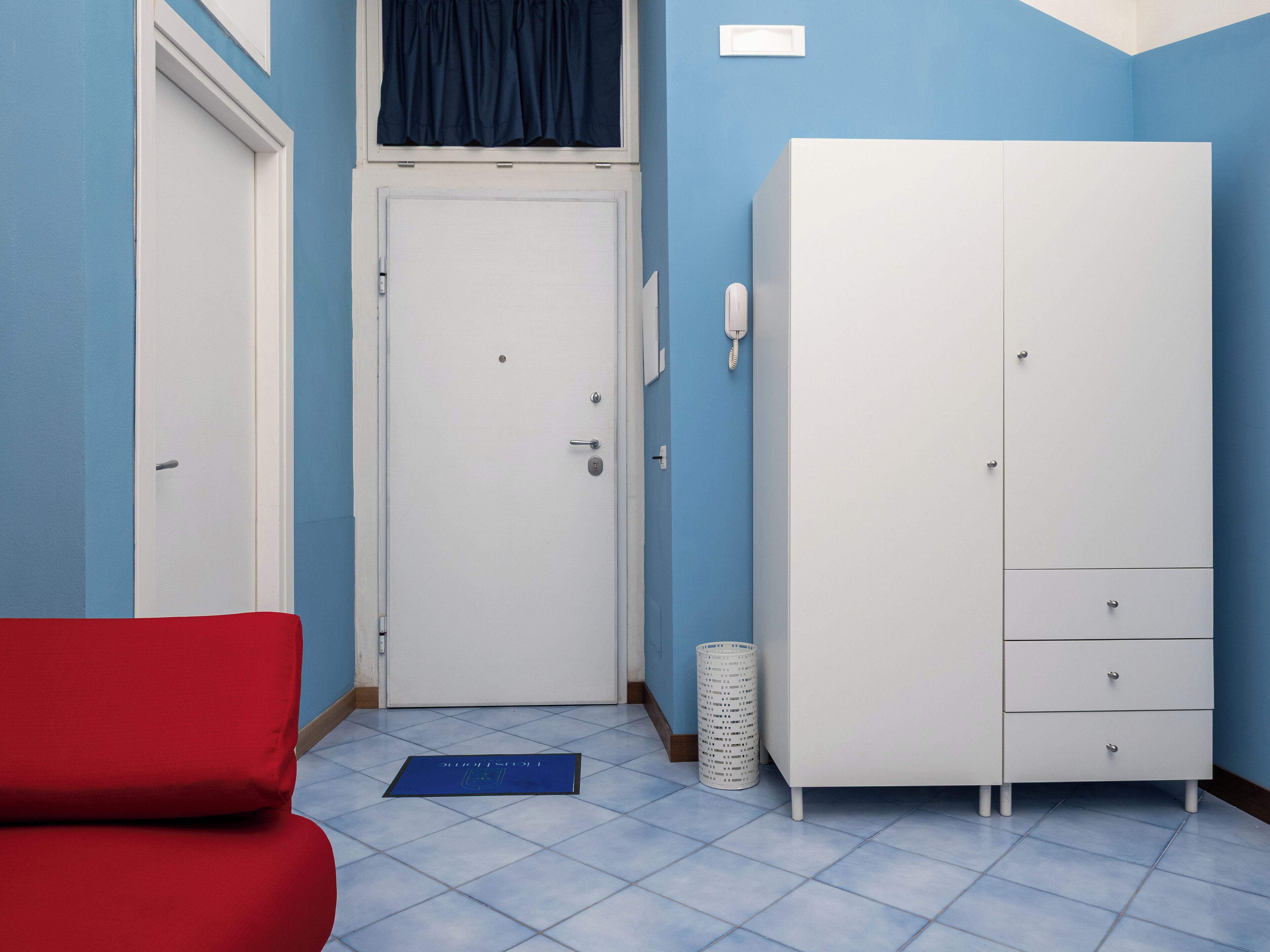 A room with a white wardrobe, a red chair, a blue mat, and a white trash bin.