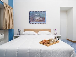 Een hotelkamer met een groot bed, wit beddengoed, een schilderij aan de muur, een dienblad met gebak en kopjes, en een bureau met een lamp.
