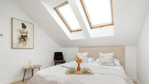 A bedroom with a bed, a bedside table, a chair, a skylight, and a painting on the wall.