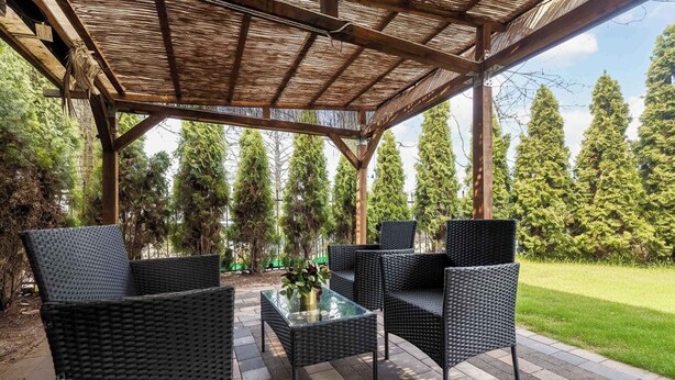 An outdoor patio with wicker furniture and a glass table, surrounded by a wooden pergola and a grassy area with trees.