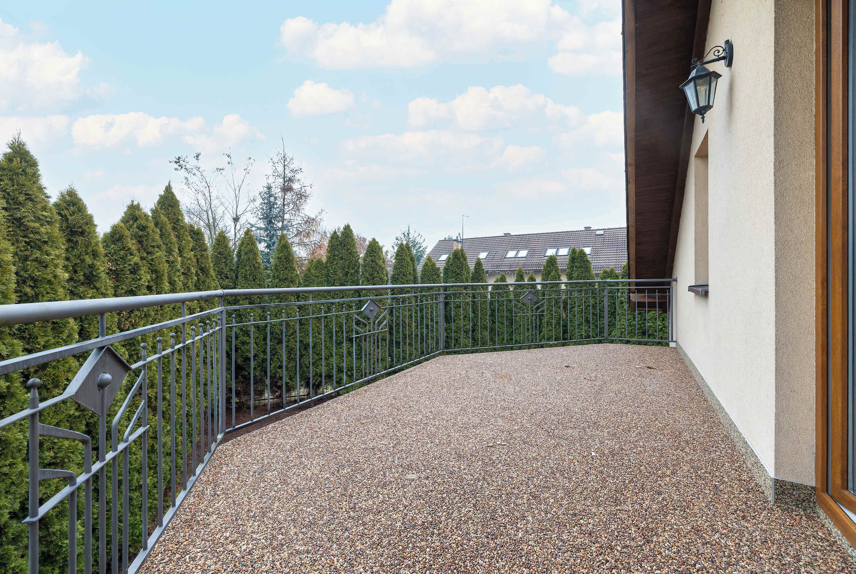 Een balkon met een grindvloer, een metalen hekwerk en uitzicht op bomen en een gebouw op de achtergrond.