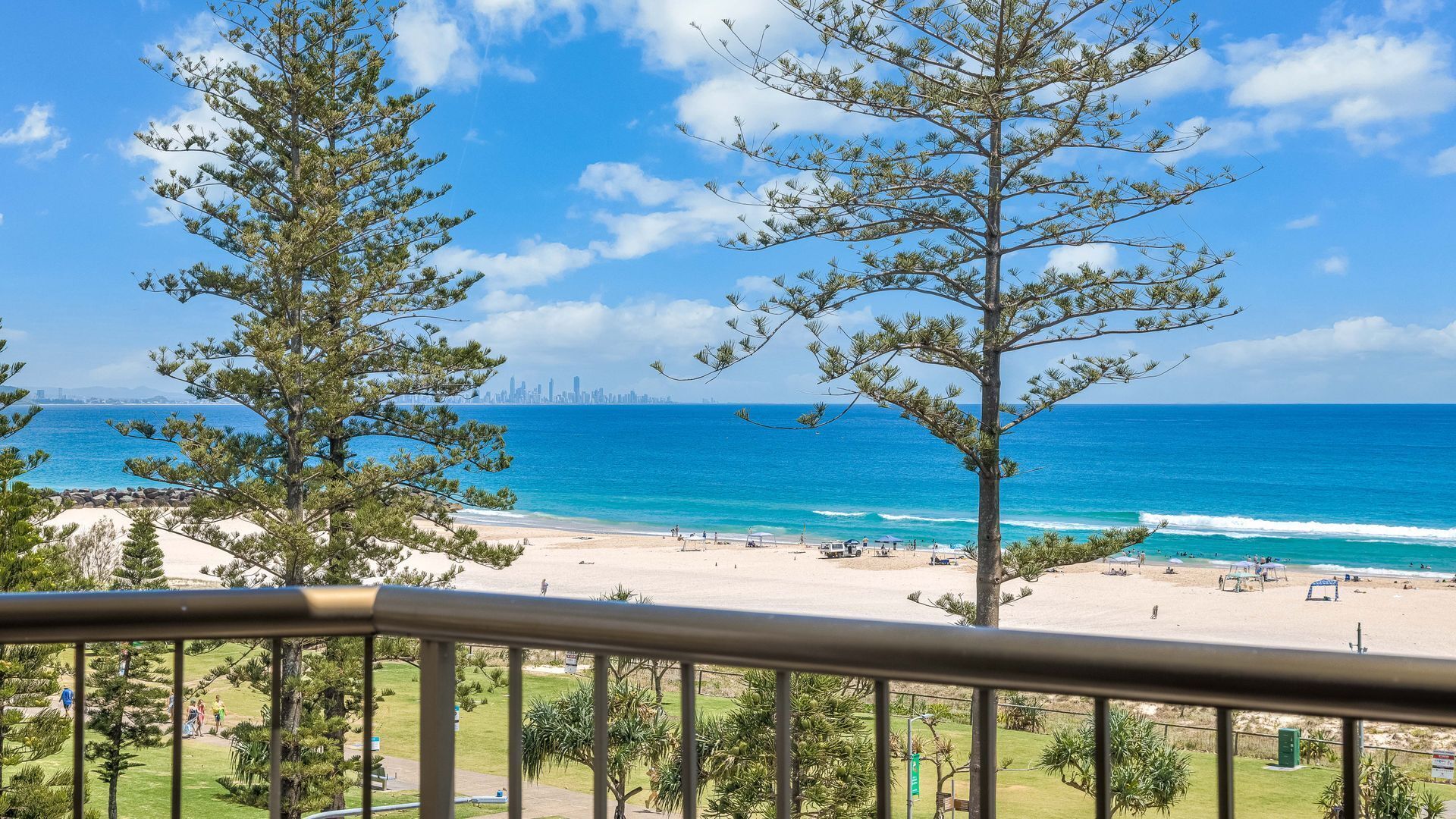 A beach view with a city skyline in the distance, trees in the foreground, and a clear blue sky.