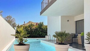 Un espace piscine extérieur moderne, agrémenté de plantes en pot, d’une terrasse en bois et offrant une vue sur un bâtiment en arrière-plan.