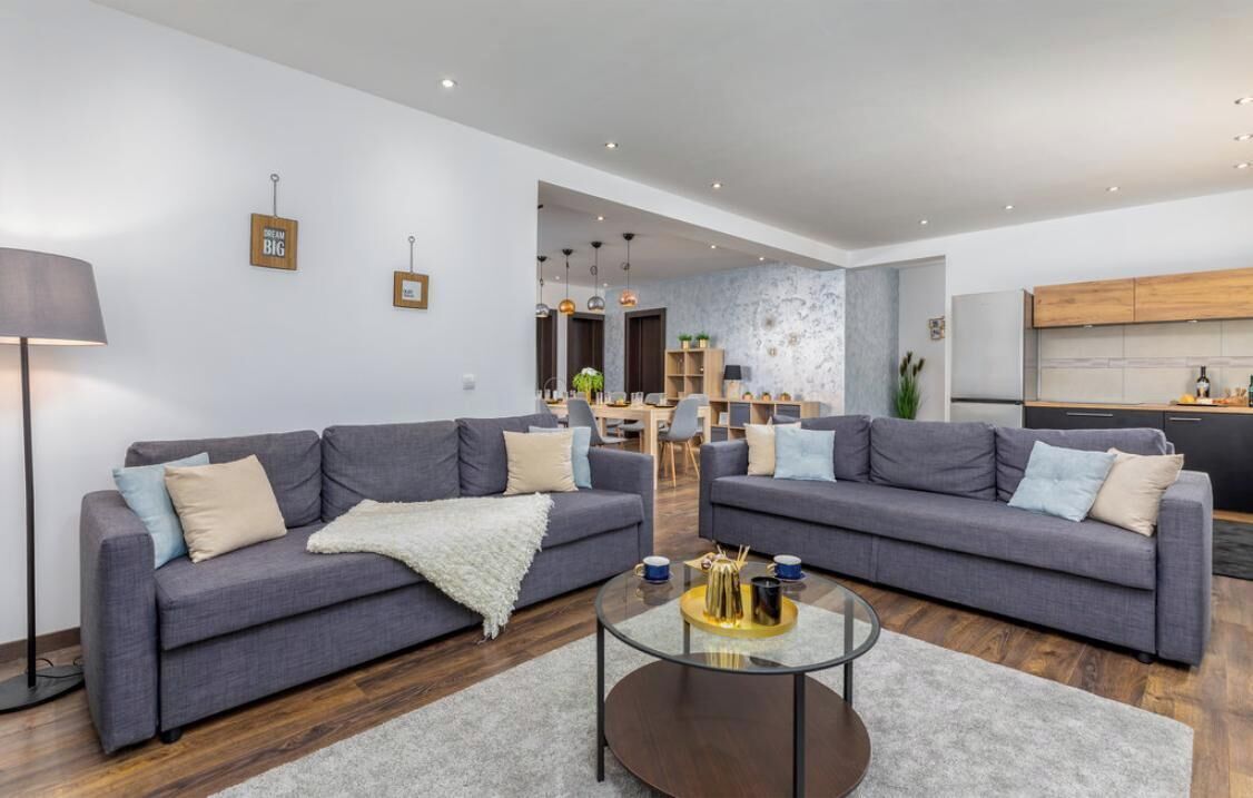A modern living room with two grey sofas, a glass coffee table, and a kitchen area in the background.