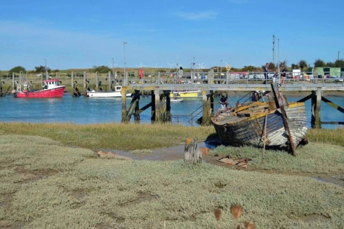 A coastal scene with a dock, a red boat, and a large, rusted shipwreck on the shore.