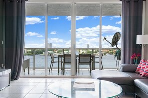 A modern living room with a glass coffee table, a sofa, and a view of a body of water through large sliding doors. - Beautiful Bay View · Walk to Sunny Isles Beach (Sunny Isles Beach)