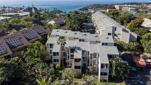 A modern multi-story building with solar panels on the roof, surrounded by lush greenery and a view of the ocean. - Kailua Bay Resort#4-204: Heart of Downtown, Walk to everything! (Kailua Kona)