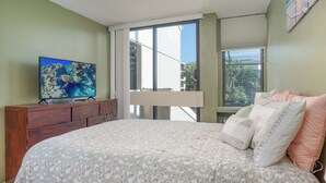 A bedroom with a bed, a TV on a wooden dresser, and a window with a view of greenery. - Kailua Bay Resort#4-204: Heart of Downtown, Walk to everything! (Kailua Kona)