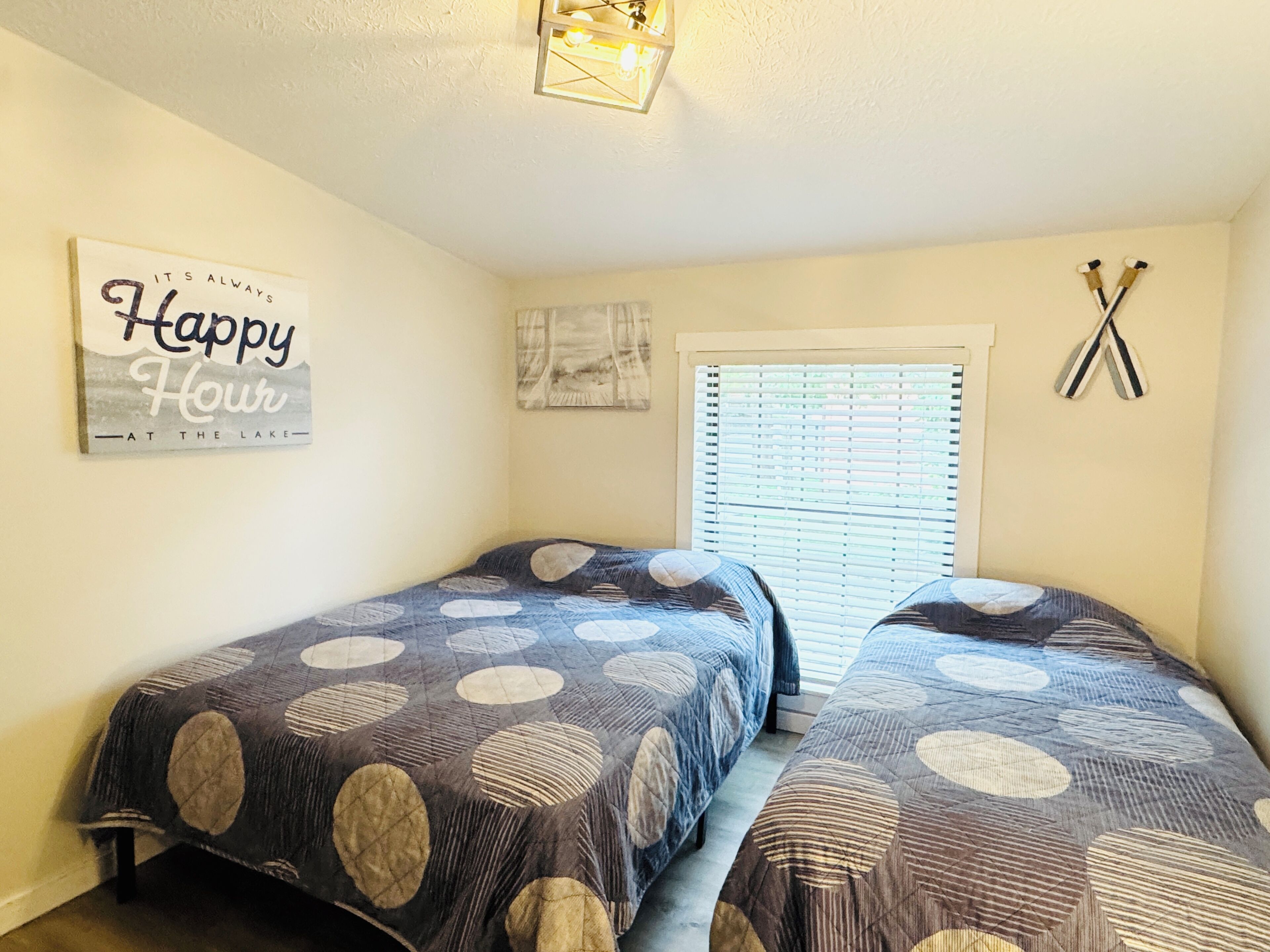 Une chambre avec deux lits, une fenêtre avec des stores, un luminaire fixé au mur et une pancarte indiquant : « Happy Hour au bord du lac ».