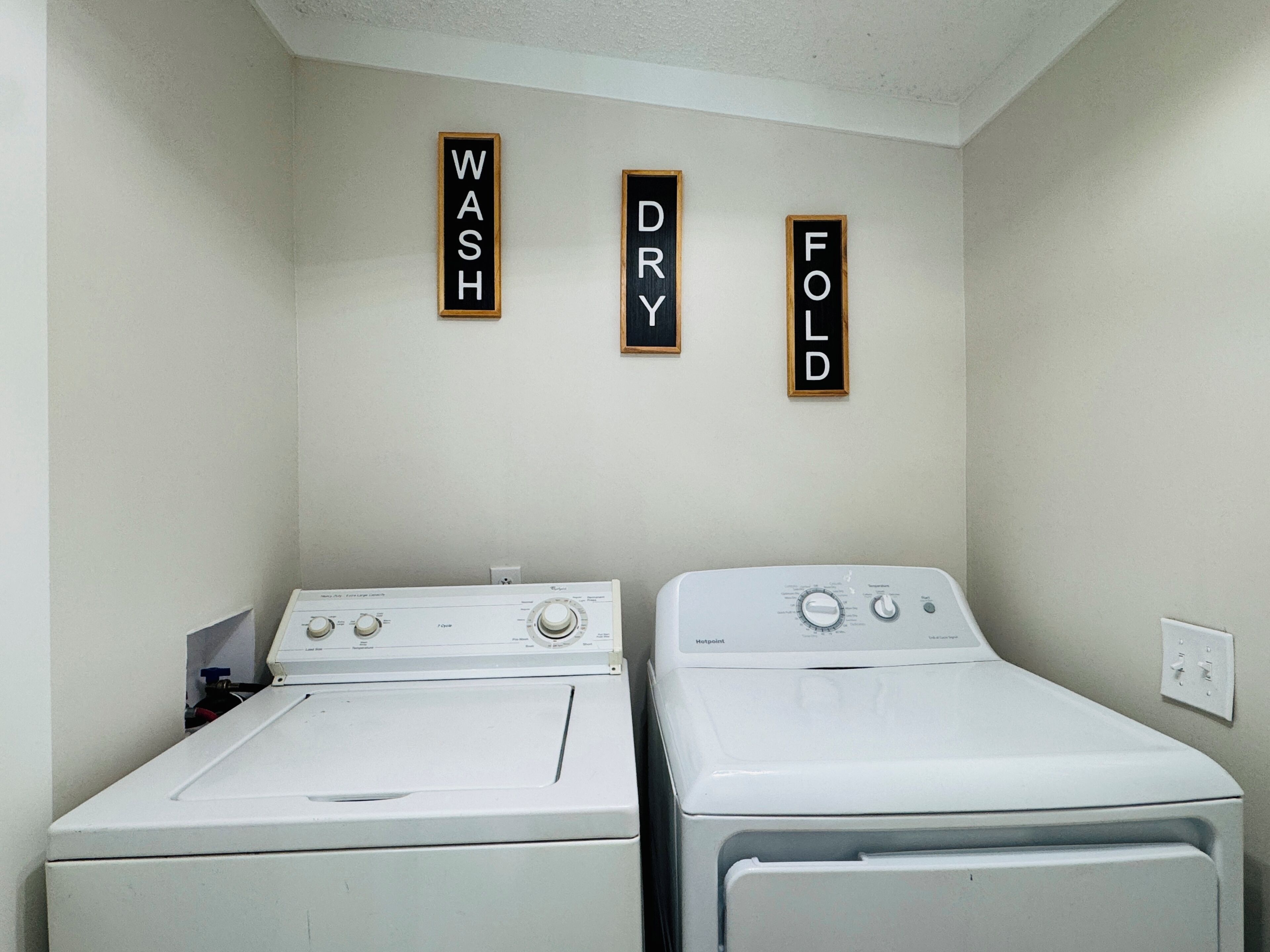 Une buanderie avec deux machines à laver blanches et trois panneaux en bois fixés au mur, portant les inscriptions « LAVAGE », « SÉCHAGE » et « PLIAGE ».